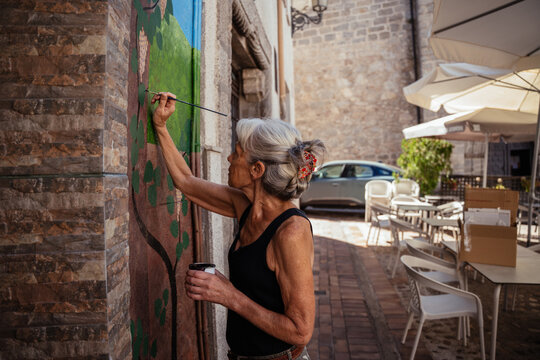 Active Senior Woman Painting Outdoor Wall Mural with Fine Brush on Sunny Street in Spanish Town