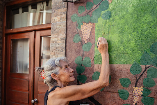 Senior Woman Carefully Painting Details on Colorful Outdoor Mural with Grape Vine Design