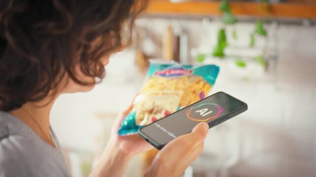 A woman in the kitchen holds a pasta package and talks to an AI voice assistant on her smartphone, studying the product ingredients. Concept of modern technology and smart home cooking
