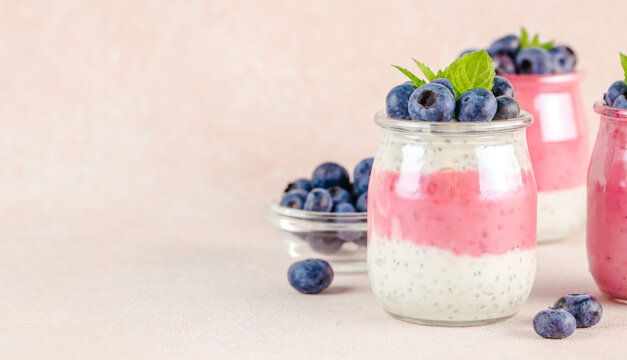 Layered yogurt dessert in glass jars with pink and white layers topped with fresh blueberries and mint leaves on a light pastel background. copy space.