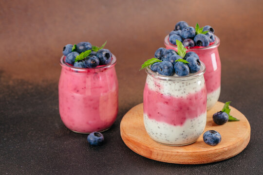 Three jars of layered yogurt with blueberries and mint leaves on a wooden board against a dark background, showcasing colorful dessert presentation
