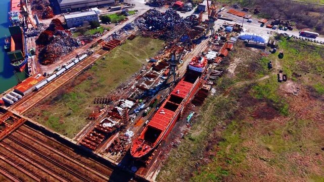 Aerial view of shipyard complex with vessels and scrapyard. Wide shot captures dry dock ships, rail tracks, warehouse and metal scrap piles.
