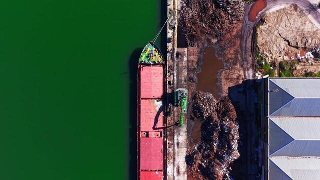 Cargo ship deck beside scrap pile, port. Directly above shot shows red hatch covers, green water and rusted metal heap onshore.