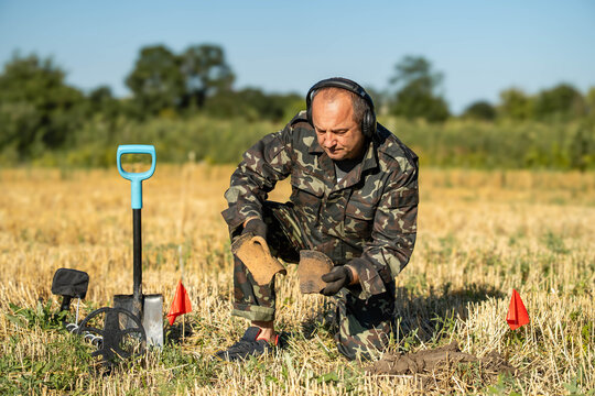 A treasure hunter is kneeling in a field with a metal detector. He is dressed in a military uniform and has headphones on. Small flags mark areas of interest around him