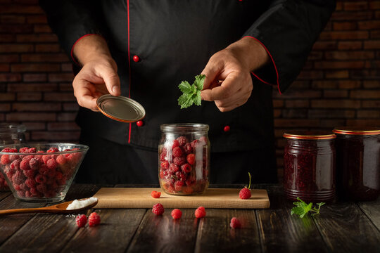 A person in a black chef jacket is adding mint leaves to a jar filled with raspberries. A bowl of raspberries and empty jars are on the wooden table in a cozy kitchen