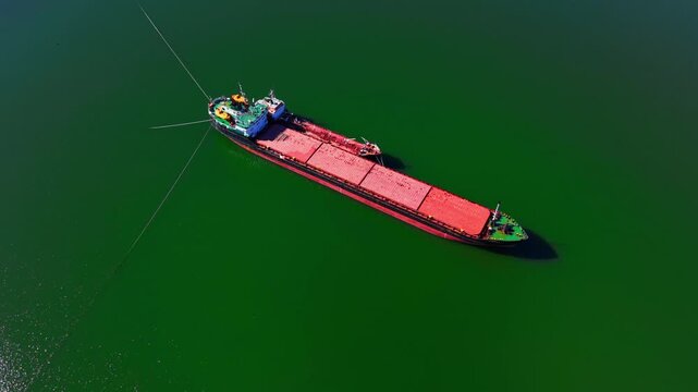 Overhead view of cargo ship anchored on green water. Directly above shot shows red hatch covers, tugboat astern and rope lines trailing.