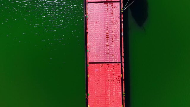 Overhead view of cargo ship deck on green water. Directly above shot captures red hatch covers, weathered steel plating and mooring ropes.
