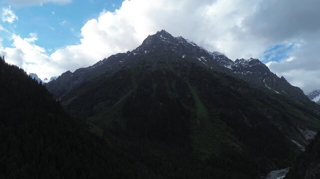 Static 4K drone footage smoothly approaches the foot of a mountain in Tekhuri gorge, Georgia. Forested hillsides lead to a rocky, snow-capped peak with clouds under a blue sky.