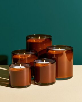 Amber glass candle vessels arranged in graduated sizes with lit flames against teal background
