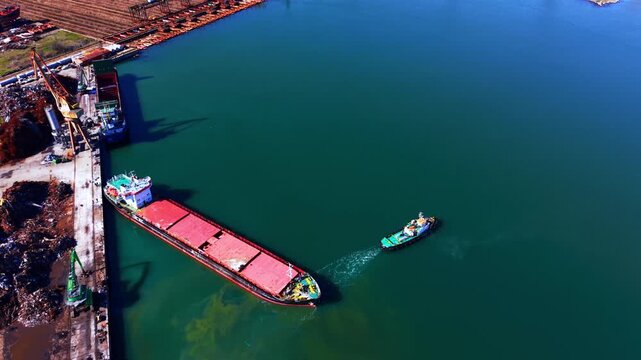 Aerial shot of tugboat pulling cargo ship, Burgas, Bulgaria. Drone view of a red bulk carrier being towed past a scrapyard along a green harbor channel.