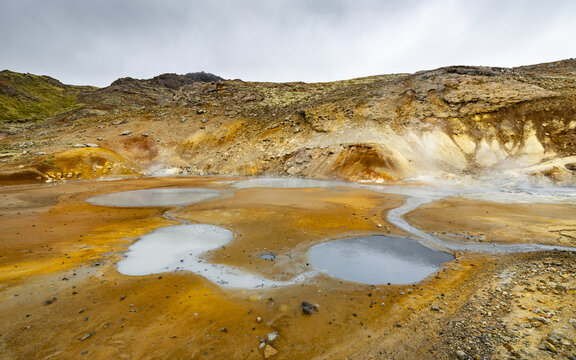 View of Seltun Geothermal Area with bubbling mud pots, steaming vents, and vibrant mineral-rich soil under a cloudy sky in Grindavik, Hafnarfjardarkaupstad, Iceland.