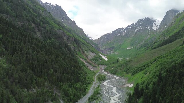 Static 4K drone footage flies smoothly backward in the Tekhuri gorge, Georgia, pulling away to reveal a wide panorama of forested foothills, snow-capped peaks, and an overcast sky.