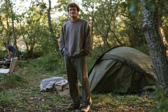A smiling young man stands confidently before his camping tent, embracing the peaceful wilderness and evoking feelings of joy and relaxation in nature's embrace.