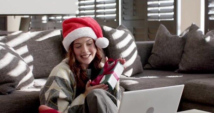20s woman sitting on gray sofa showing wrapped gift with red ribbon to laptop during call