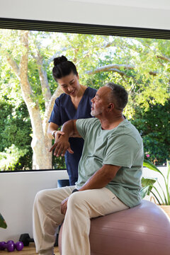 Asian caregiver in navy scrubs guiding senior African American man on pink ball in rehab room