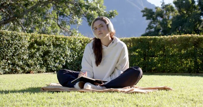 Hoodie-clad woman sitting on blanket on lawn, listening on headset and writing in notebook with pen