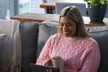 Mid adult African woman in pink sweater sitting on sofa at home holding tablet and mug © WavebreakMediaMicro
