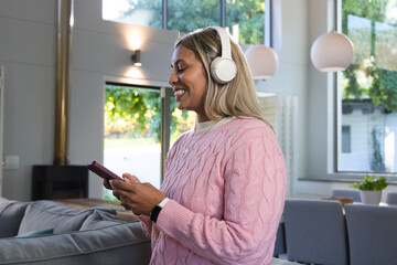 Naklejka premium Woman wearing pink sweater and white headset standing by sofa holding phone, smiling in living room
