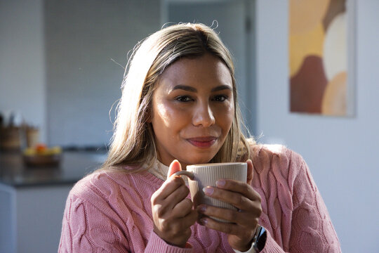 African American woman holding ribbed mug close to face in pink sweater with watch in kitchen