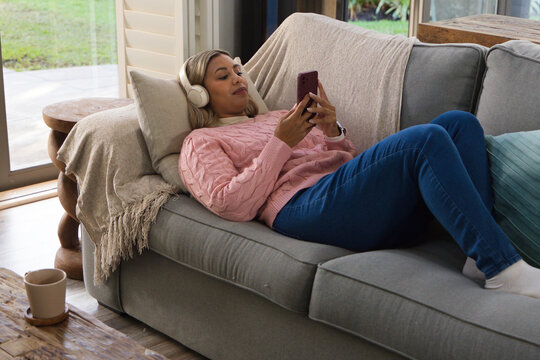 Mid-adult African American woman reclining on sofa at home in sweater headphones holding red phone
