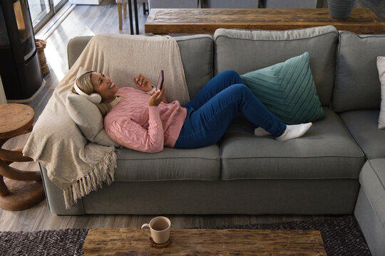 African woman lying across gray sofa at home, wearing pink sweater, headphones, holding smartphone