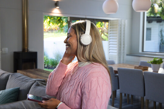 African woman wearing pink sweater listening with white headset holding smartphone at home