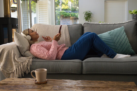 African American woman lying on sofa at home in pink sweater wearing headset and holding phone