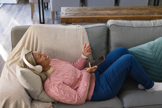 African American woman reclining on gray sofa at home, wearing headphones and holding smartphone