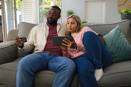 Diverse couple sitting on gray sofa in bright living room holding tablet and credit card
