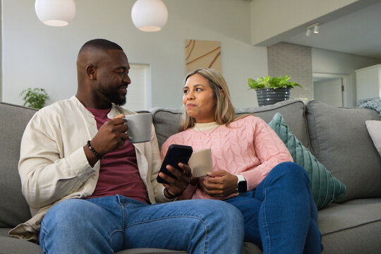 Diverse couple sitting together on gray sofa holding ceramic mugs and smartphone in living room