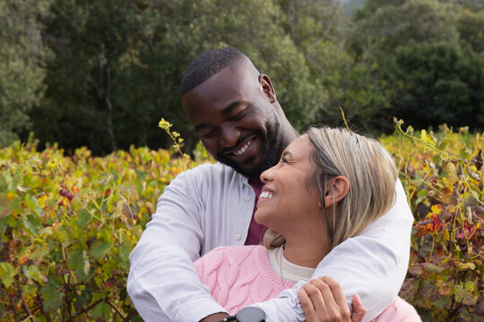 Diverse couple embracing among vineyard rows during autumn, man wearing smartwatch and button-up