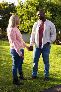 Diverse couple holding hands and smiling while standing on sunlit backyard lawn showing wristwatch