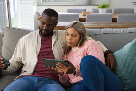 African American couple sitting on grey sofa at home, using tablet while holding credit card