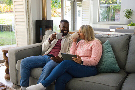 Diverse couple sitting on gray sofa near sliding doors, holding mugs, using tablet and smartphone