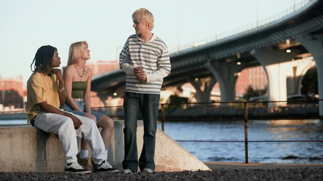 Multiethnic teenagers hanging out by riverbank near bridge, talking and gesturing. Footage is suitable for youth lifestyle marketing, diversity campaigns, and social connection advertisements