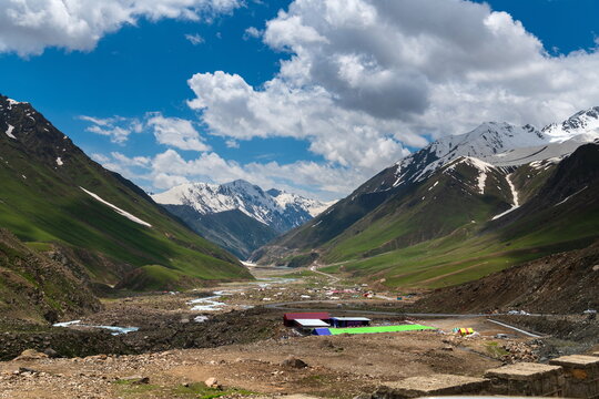 View of Gittidas valley with lush green slopes, snow-capped mountain peaks, a winding road, and a small settlement under a blue sky with clouds in Gittidas, Khyber Pakhtunkhwa, Pakistan.