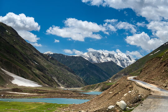 View of the Gittidas valley with a winding road, a calm river, and snow-capped mountains under a blue sky in Gittidas, Khyber Pakhtunkhwa, Pakistan.