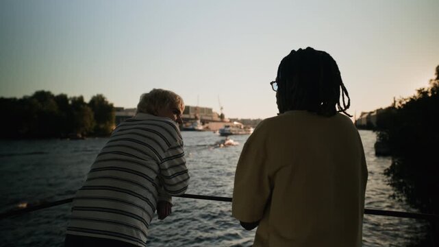 Caucasian young woman and Black young man standing by railing, watching boats on river at sunset. Footage is suitable for diversity, friendship, and urban lifestyle promotional videos