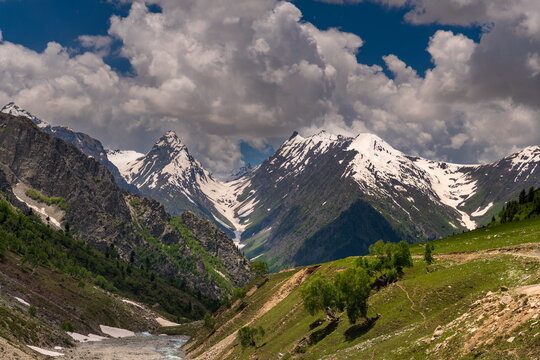 View of Minimarg Astore featuring snow-capped mountain peaks, lush green slopes, and a flowing stream under a dramatic cloudy sky in District Astore, Gilgit Baltistan, Pakistan.