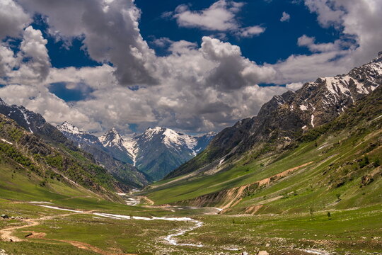 View of the lush green valley of Minimarg Astore with snow-capped mountain peaks, a winding stream, and dramatic clouds in the sky in District Astore, Gilgit Baltistan, Pakistan.