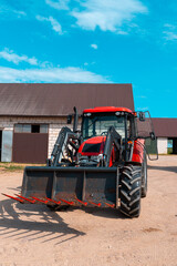 Red farm tractor with front loader isolated in a rural farmyard, modern agricultural machinery parked near barn buildings under a bright blue sky © Iryna