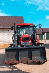 Red farm tractor with front loader isolated in a rural farmyard, modern agricultural machinery parked near barn buildings under a bright blue sky © Iryna