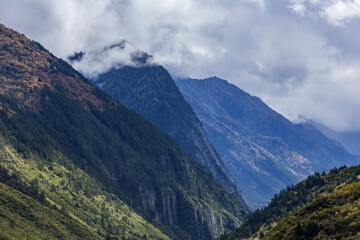 Layered mountain peaks, forested slopes and misty clouds form a natural landscape in a valley under an overcast sky.