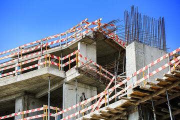 Fototapeta premium Construction site with reinforced concrete structure, rebar columns and striped safety barrier tape under clear sky, building renovation progress