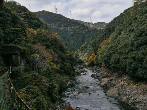 View of the Hozugawa River flowing through the forested Arashiyama valley with a railway tunnel and tracks on the left in Kyoto, Kyoto, Japan.