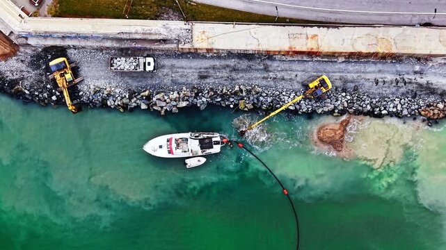 Two excavators dredging harbor edge. Top down drone shot of yellow machinery, boats and a hose stretching across green water.