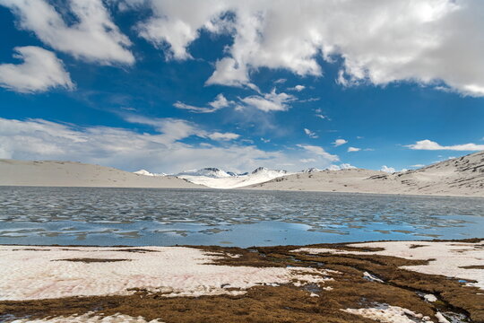 View of Sheosar Lake with its partially frozen surface, surrounded by snow-capped mountains under a bright blue sky with scattered clouds in Skardu, Gilgit Baltistan, Pakistan.