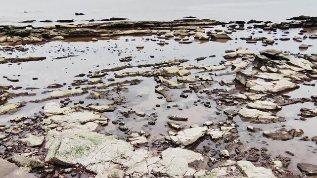 Wide shot panning motion video of a rocky shoreline with uneven flat stones partially submerged in calm shallow water. The textured stone formations lead toward the horizon