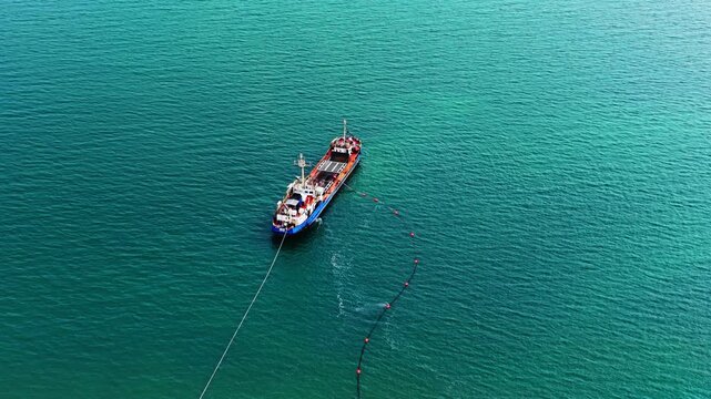 Above view of vessel towing floating boom, Black Sea coast. Blue and red ship pulls a buoyed containment line across calm turquoise water.