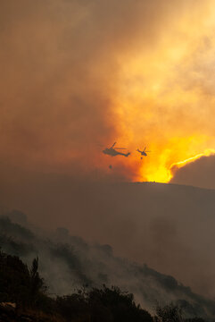 View of helicopters fighting a wildfire by dropping water over a smoke-filled hillside at sunset in South Africa.